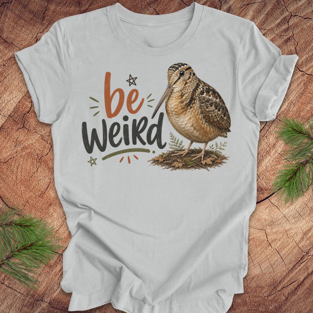 A light gray graphic t-shirt featuring the text 'be Weird' in orange and dark gray, alongside an illustration of an american woodcock bird. The t-shirt is displayed flat on a rustic wood background decorated with pine branches.
