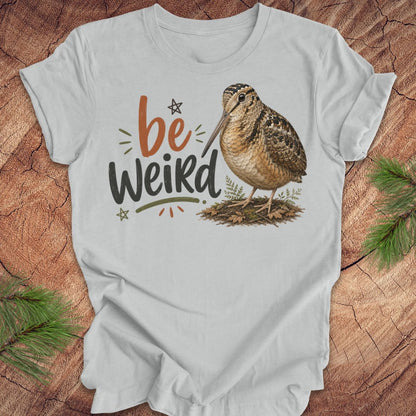 A light gray graphic t-shirt featuring the text 'be Weird' in orange and dark gray, alongside an illustration of an american woodcock bird. The t-shirt is displayed flat on a rustic wood background decorated with pine branches.