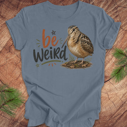 A slate blue graphic t-shirt featuring the text 'be Weird' in orange and dark gray, alongside an illustration of an american woodcock bird. The t-shirt is displayed flat on a rustic wood background decorated with pine branches.