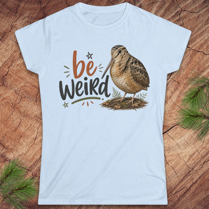 A light blue crewneck t-shirt featuring a graphic print of a brown woodcock bird and the text 'be Weird' in stylized lettering, displayed flat on a rustic wood background with pine branches.