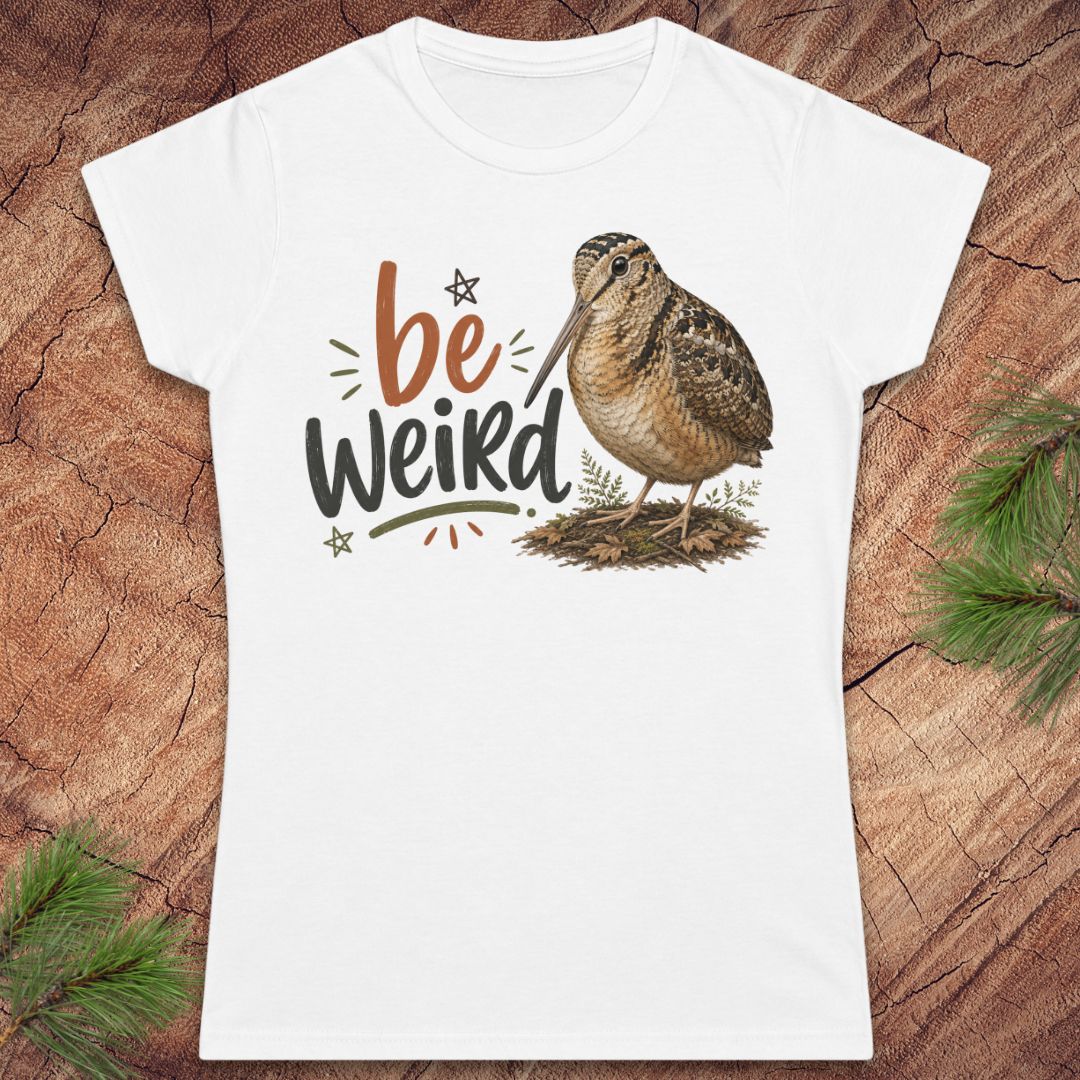 A white women's short-sleeve t-shirt with a graphic print of a brown woodcock and the text 'be Weird' in brown and black, displayed flat on a rustic wood background with pine branches.