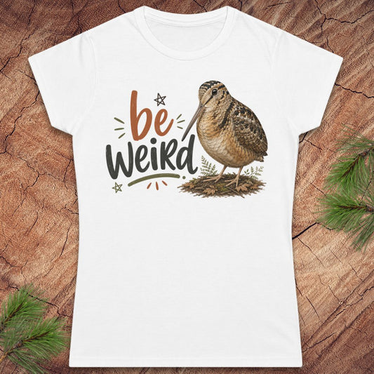 A white women's short-sleeve t-shirt with a graphic print of a brown woodcock and the text 'be Weird' in brown and black, displayed flat on a rustic wood background with pine branches.