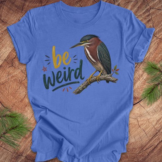 A blue short-sleeve crewneck t-shirt laid flat on a rustic wood background with pine branches in the corners. The shirt features a graphic of a green heron perched on a branch next to the hand-lettered text 'be weird' in yellow and dark green.