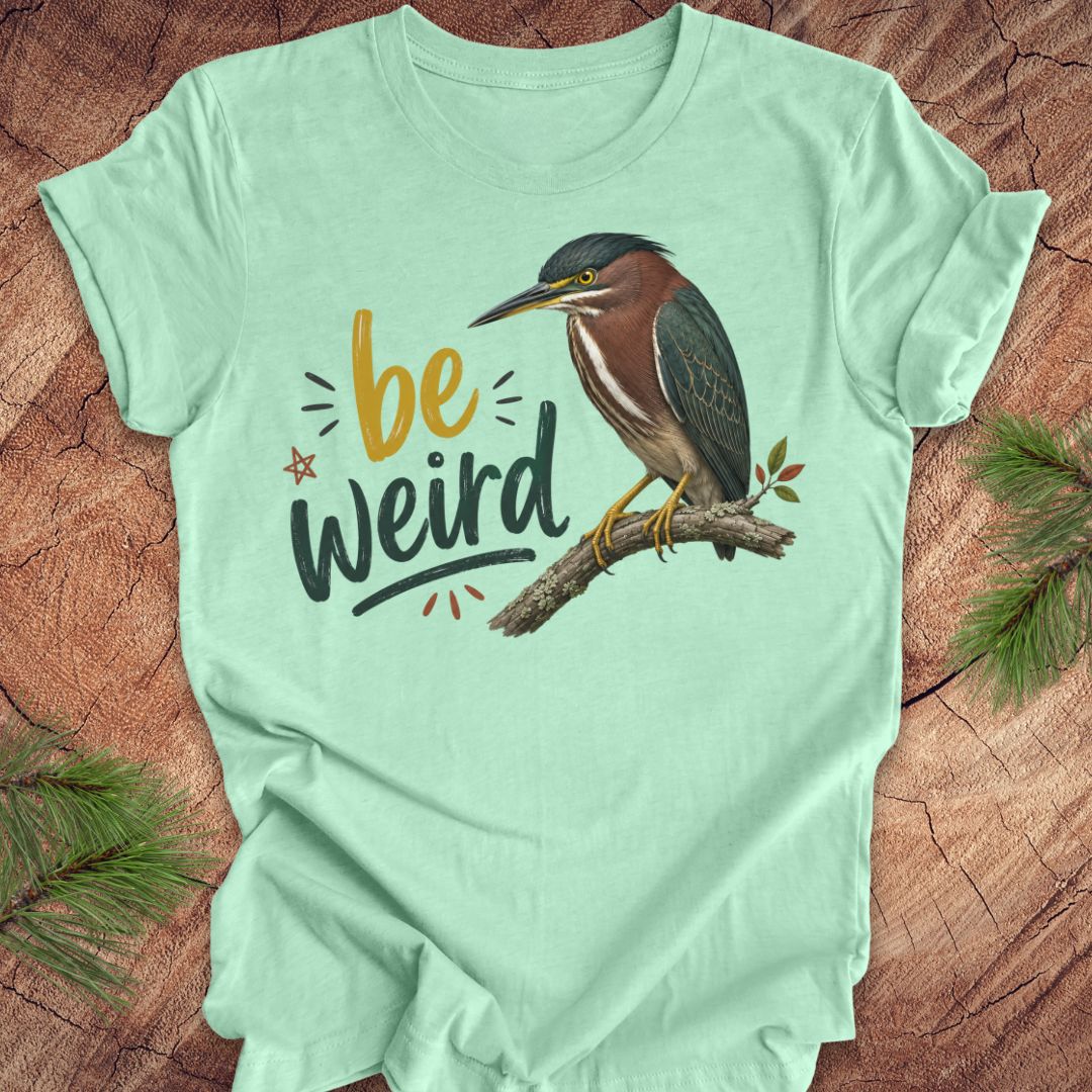 A mint green short-sleeve t-shirt laid flat on a rustic wood background with pine branches in the corners. The shirt features a graphic of a green heron perched on a branch next to the text 'be weird' in yellow, black, and gray lettering.