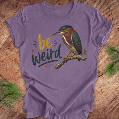 A purple short-sleeve t-shirt with a graphic print of a green heron bird perched on a branch next to the text 'be weird' in yellow and dark green lettering. The shirt is displayed flat on a rustic wood background with pine branches in the corners.