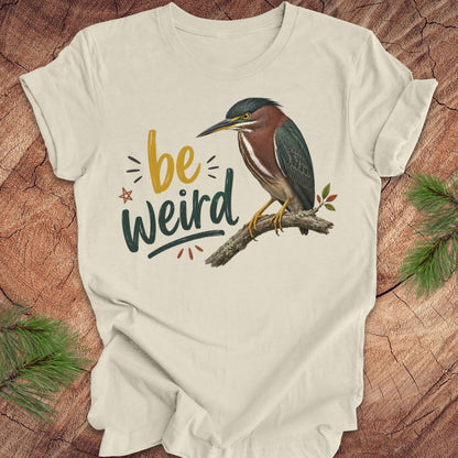 An off-white short-sleeve t-shirt laid flat on a rustic wood background, featuring a graphic of a green heron perched on a branch with the text 'be weird' in yellow and dark green lettering.
