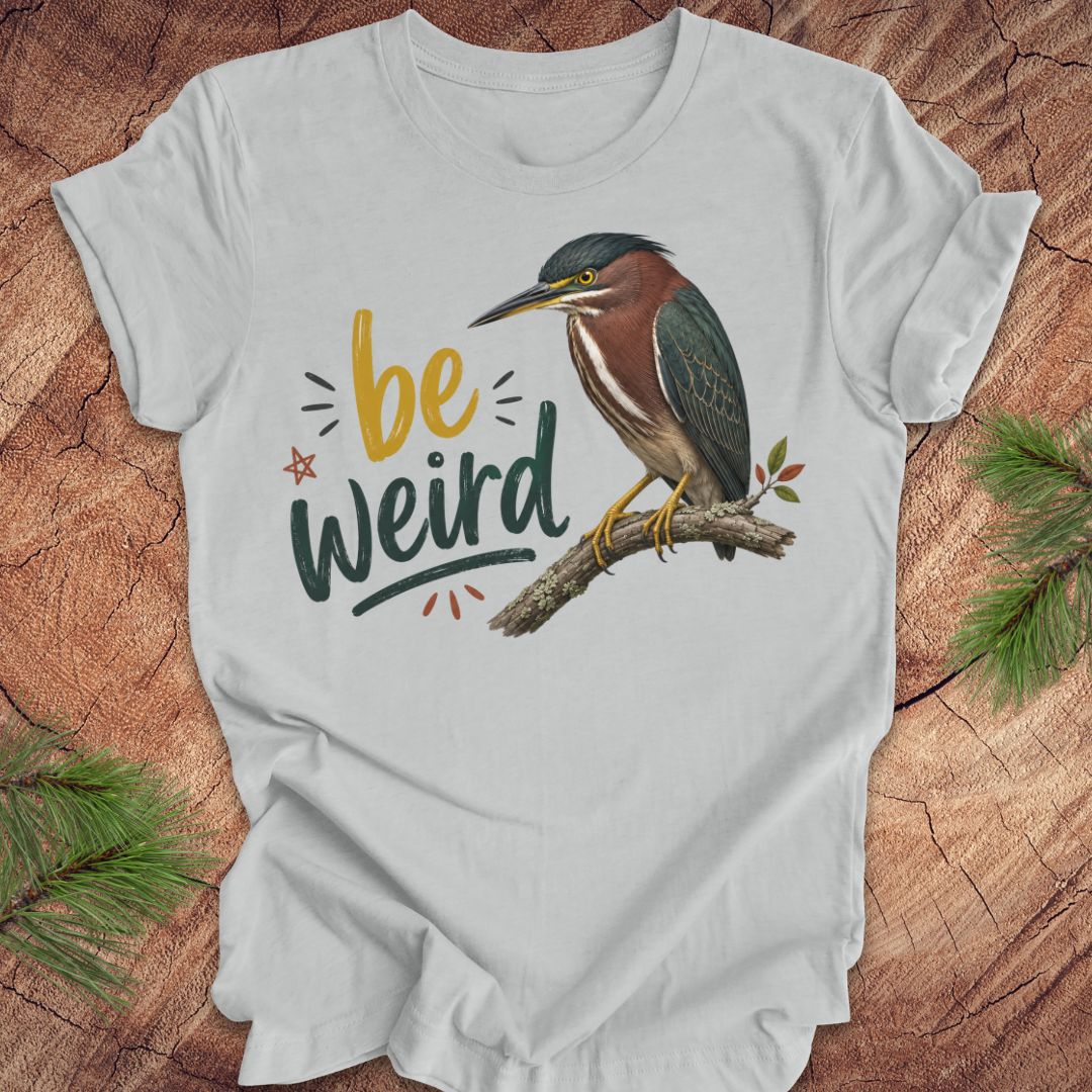 A light gray short-sleeve t-shirt laid flat on a rustic wood background with pine branches in the corners. The shirt features a graphic of a green heron perched on a branch next to the text 'be weird' in yellow and dark green lettering.