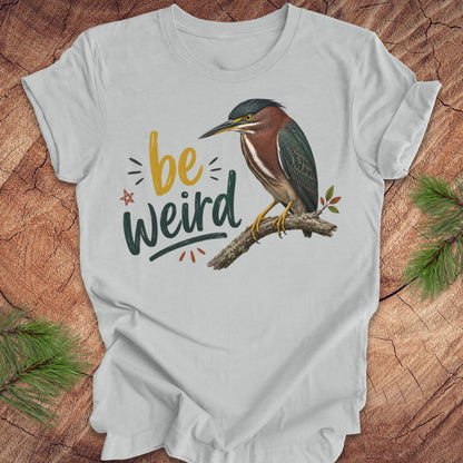 A light gray short-sleeve t-shirt laid flat on a rustic wood background with pine branches in the corners. The shirt features a graphic of a green heron perched on a branch next to the text 'be weird' in yellow and dark green lettering.