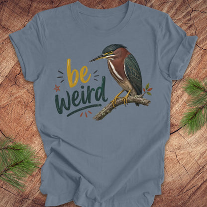 A slate blue graphic t-shirt featuring a green heron perched on a branch next to the text 'be weird' in yellow and dark green lettering. The t-shirt is displayed flat on a rustic wood background with pine needles in the corners.