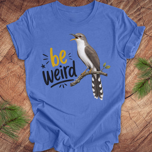 A blue short-sleeve crewneck t-shirt laid flat on a rustic wood background with pine branches in the corners. The shirt features a graphic of a yellow-billed cuckoo bird perched on a branch next to the text 'be weird' in yellow and black.