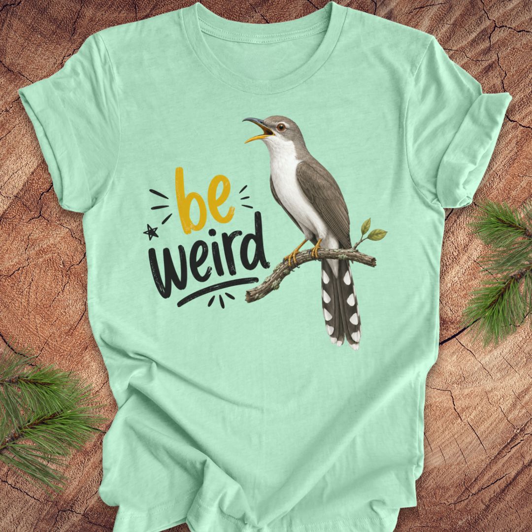 A mint green short-sleeve crewneck t-shirt laid flat on a rustic wood background with pine branches in the corners. The shirt features a graphic of a yellow-billed cuckoo bird perched on a branch next to the text 'be weird' in yellow and black.