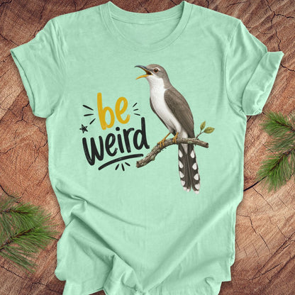 A mint green short-sleeve crewneck t-shirt laid flat on a rustic wood background with pine branches in the corners. The shirt features a graphic of a yellow-billed cuckoo bird perched on a branch next to the text 'be weird' in yellow and black.