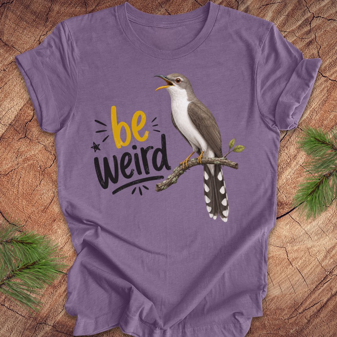 A purple short-sleeve crewneck t-shirt laid flat on a rustic wood background with pine branches in the corners. The shirt features a graphic of a yellow-billed cuckoo bird perched on a branch next to the text 'be weird' in yellow and black.
