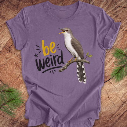A purple short-sleeve crewneck t-shirt laid flat on a rustic wood background with pine branches in the corners. The shirt features a graphic of a yellow-billed cuckoo bird perched on a branch next to the text 'be weird' in yellow and black.