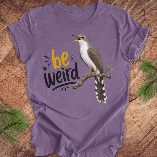 A purple short-sleeve crewneck t-shirt laid flat on a rustic wood background with pine branches in the corners. The shirt features a graphic of a yellow-billed cuckoo bird perched on a branch next to the text 'be weird' in yellow and black.