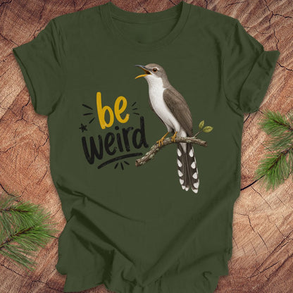 A green short-sleeve crewneck t-shirt laid flat on a rustic wood background with pine branches in the corners. The shirt features a graphic of a yellow-billed cuckoo bird perched on a branch next to the text 'be weird' in yellow and black.