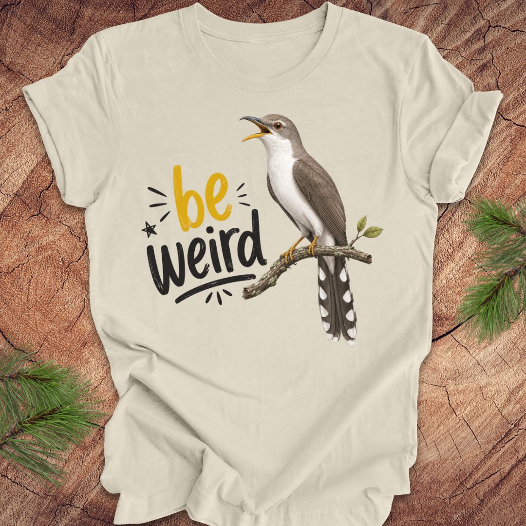 A beige short-sleeve crewneck t-shirt laid flat on a rustic wood background with pine branches in the corners. The shirt features a graphic of a yellow-billed cuckoo bird perched on a branch next to the text 'be weird' in yellow and black.