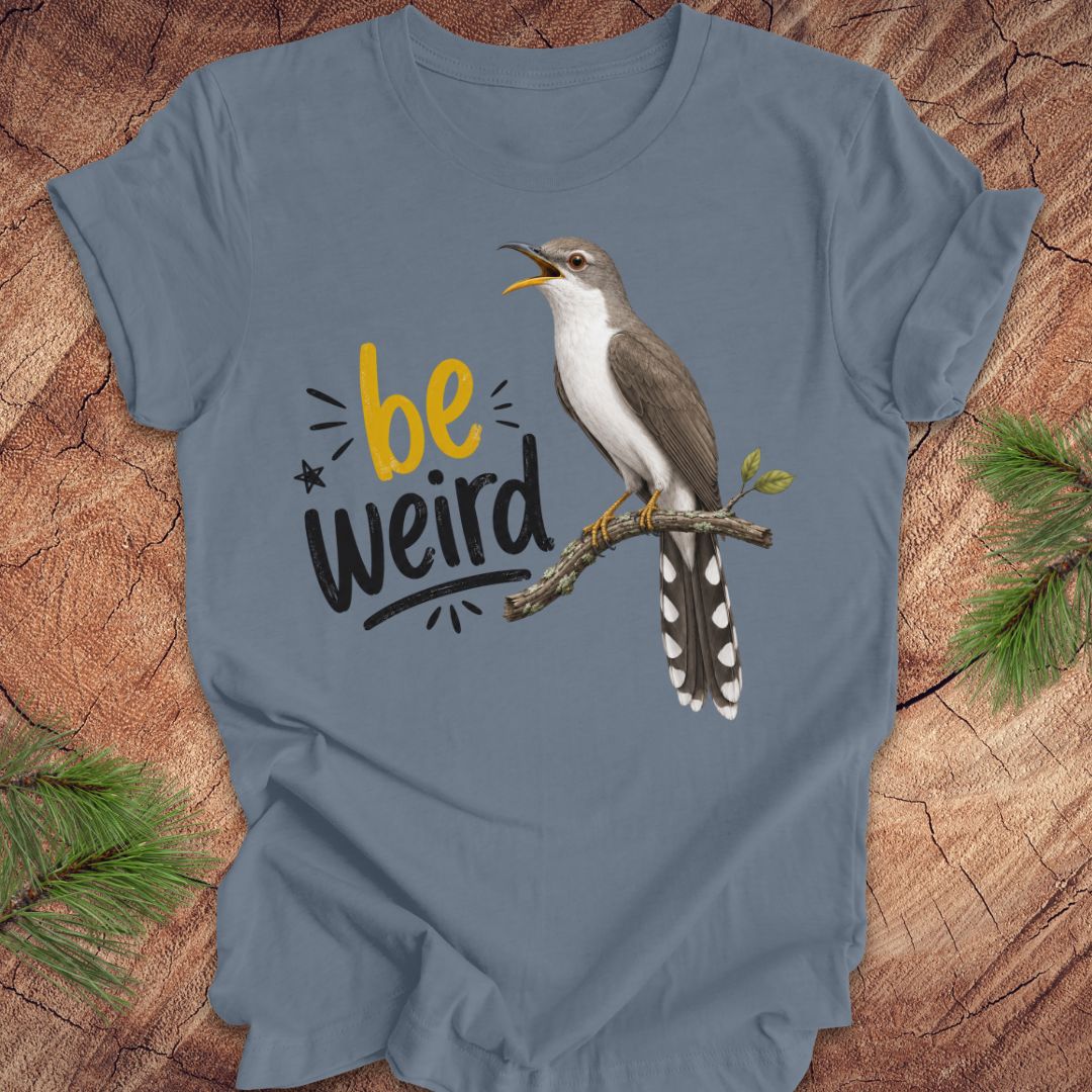 A blue short-sleeve crewneck t-shirt laid flat on a rustic wood background with pine branches in the corners. The shirt features a graphic of a yellow-billed cuckoo bird perched on a branch next to the text 'be weird' in yellow and black.