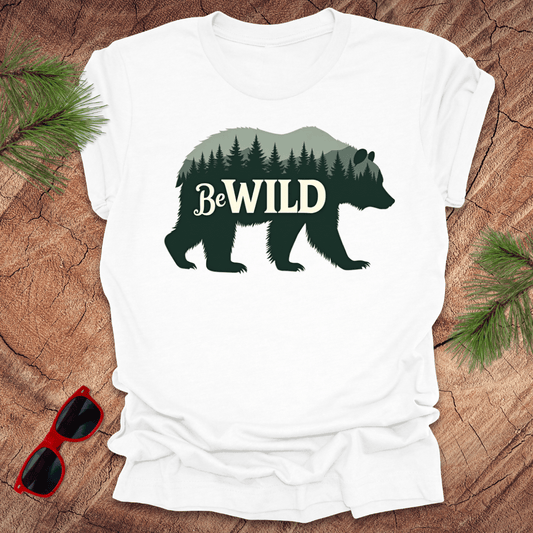 A white t-shirt with a graphic of a bear and the text 'Be Wild' on it, displayed on a wooden surface with sunglasses and pine branches.