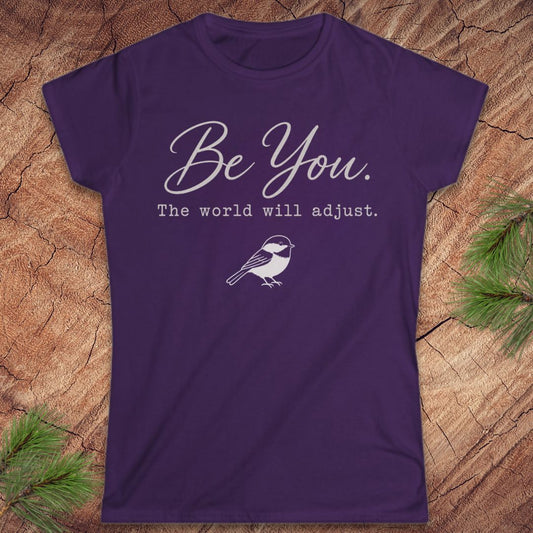 A purple short-sleeve t-shirt with a white graphic print that reads 'Be You. The world will adjust.