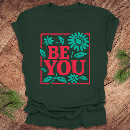 Green t-shirt with 'BE YOU' text and flowers on a wooden surface
