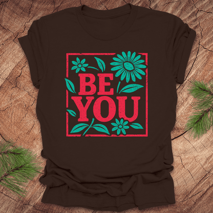Brown t-shirt with 'BE YOU' text and flowers on a wooden surface