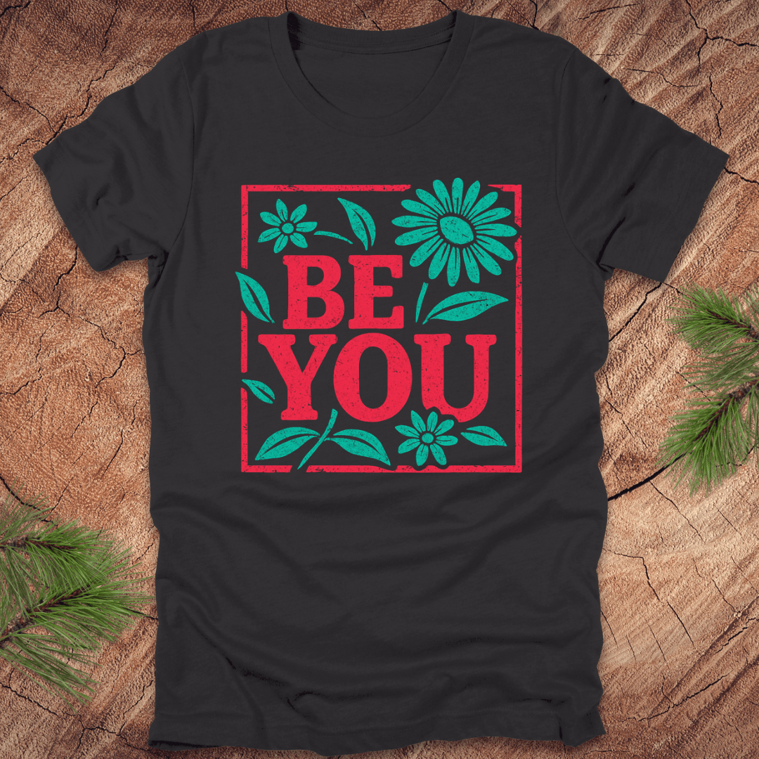 Dark grey t-shirt with 'BE YOU' text and floral design on a wooden surface