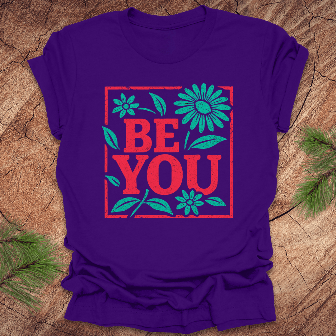 Purple t-shirt with 'BE YOU' text and floral design on a wooden surface