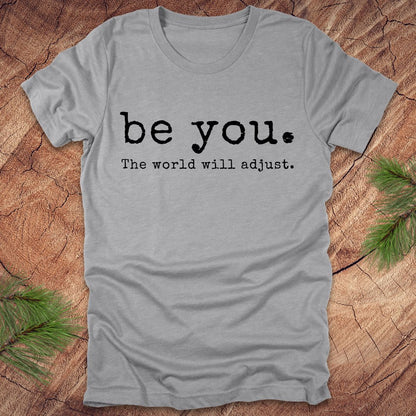 Gray t-shirt with 'be you. The world will adjust.' text on a wooden surface