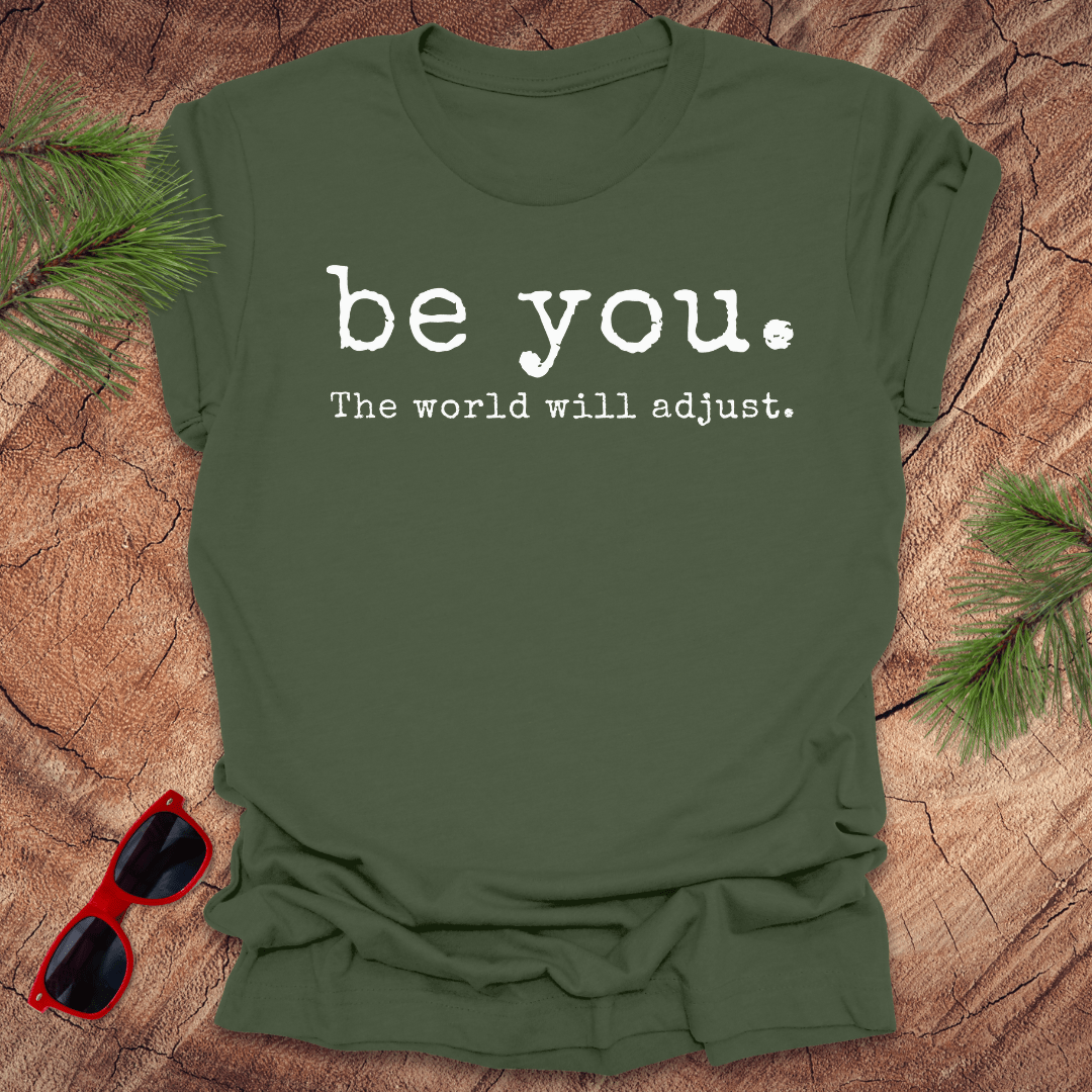 military green tshirt with the text Be you. the world will adjust on a wood background with sunglasses and pine