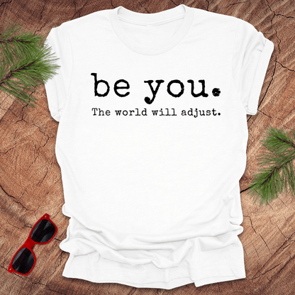 white tshirt with the text Be you. the world will adjust on a wood background with sunglasses and pine