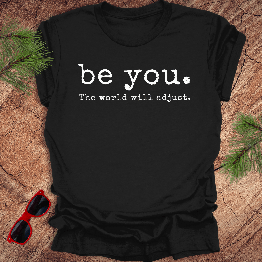 black tshirt with the text Be you. the world will adjust on a wood background with sunglasses and pine