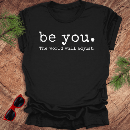 black tshirt with the text Be you. the world will adjust on a wood background with sunglasses and pine