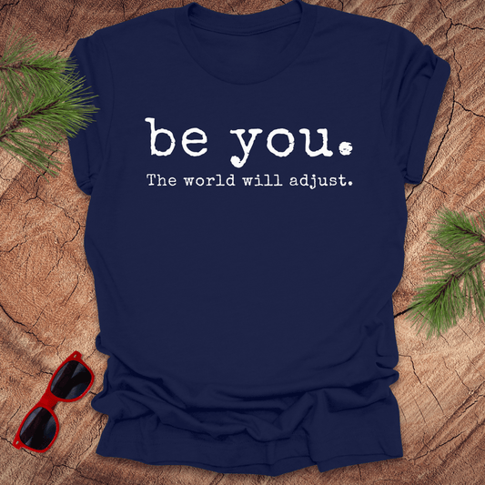 navy tshirt with the text Be you. the world will adjust on a wood background with sunglasses and pine