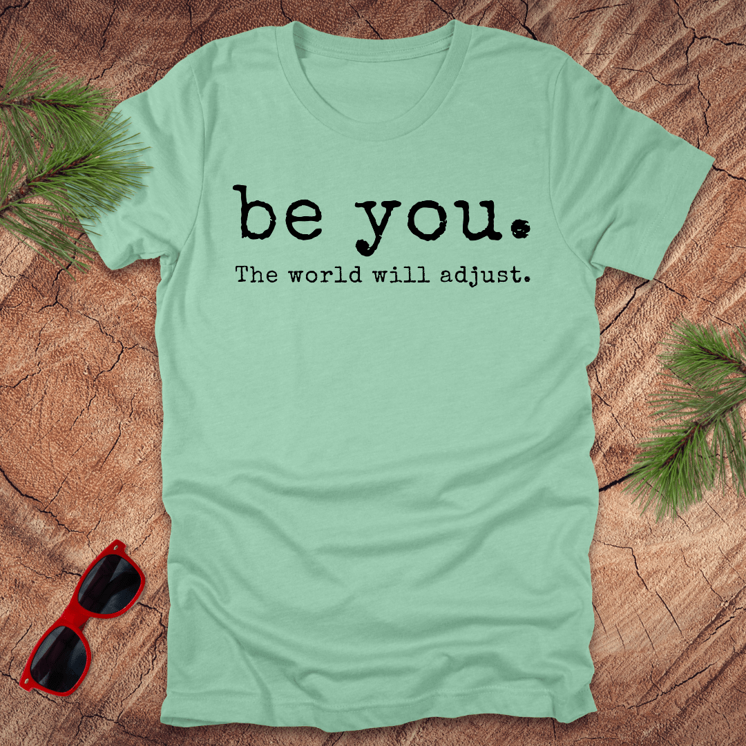 mint green tshirt with the text Be you. the world will adjust on a wood background with sunglasses and pine