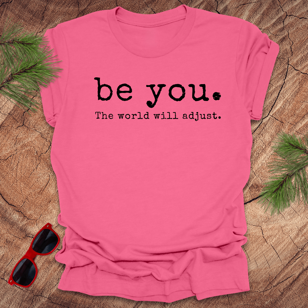 pink tshirt with the text Be you. the world will adjust on a wood background with sunglasses and pine