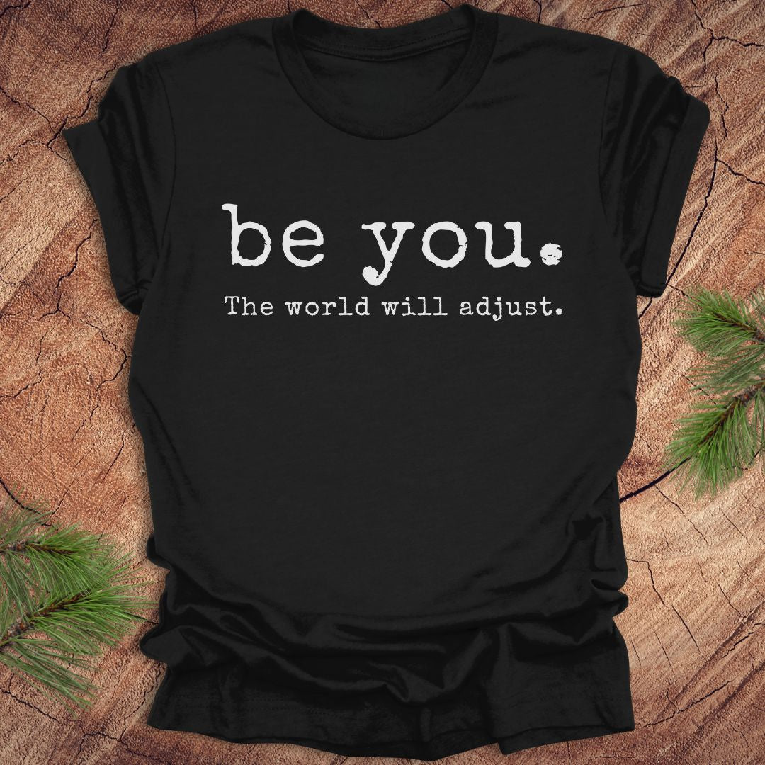 Black t-shirt with 'be you. The world will adjust.' text on a wooden surface.