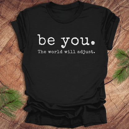 Black t-shirt with 'be you. The world will adjust.' text on a wooden surface.