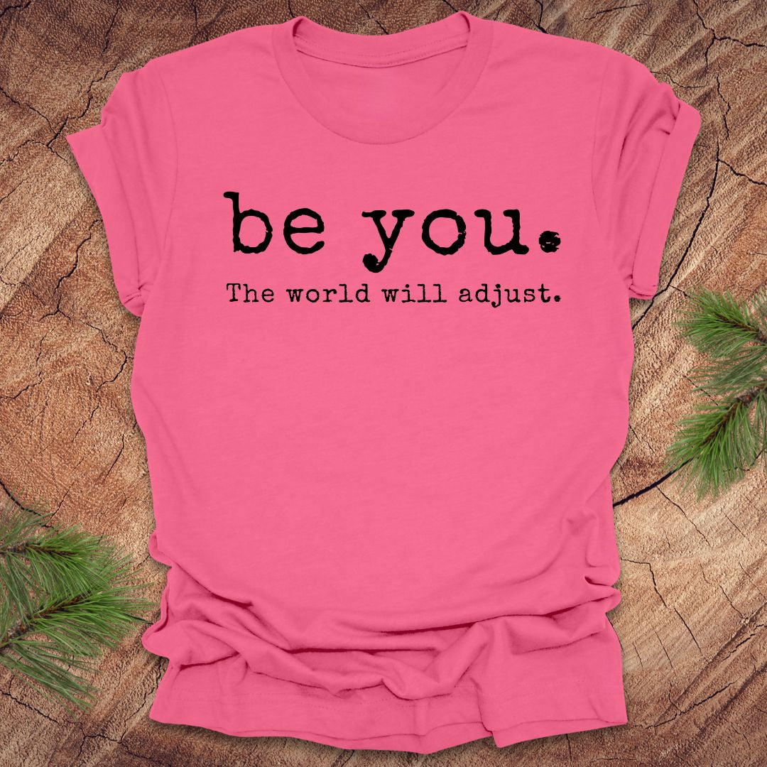 Pink t-shirt with 'be you. The world will adjust.' text on a wooden surface.