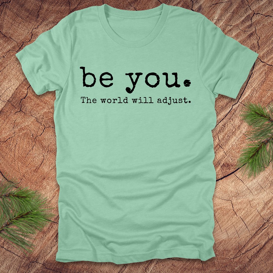 Mint green t-shirt with 'be you. The world will adjust.' text on a wooden surface.