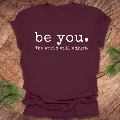 Maroon t-shirt with 'be you. The world will adjust.' text on a wooden surface.
