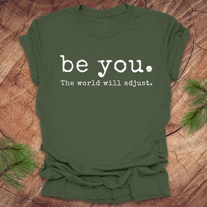 Green t-shirt with 'be you. The world will adjust.' text on a wooden surface.