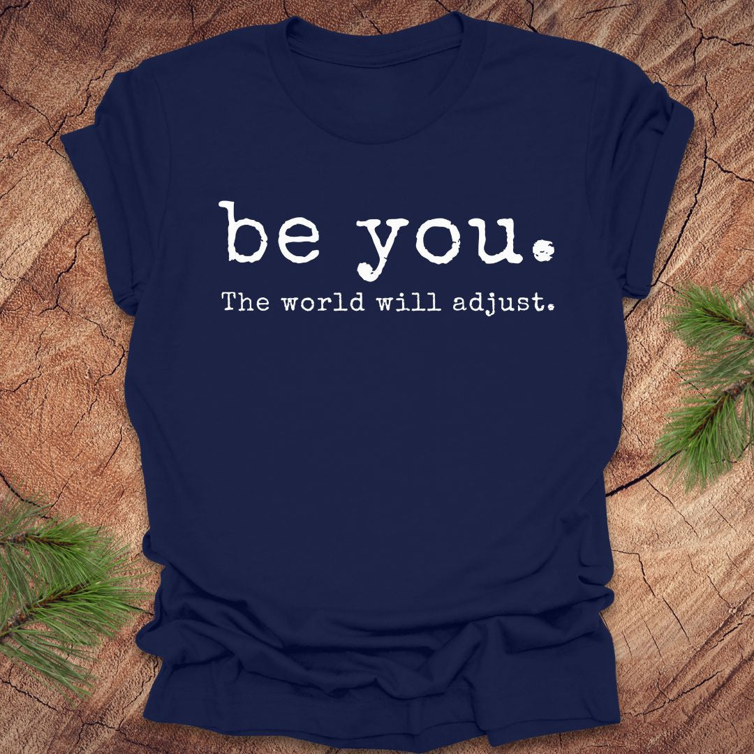 Navy blue t-shirt with 'be you. The world will adjust.' text on a wooden surface.