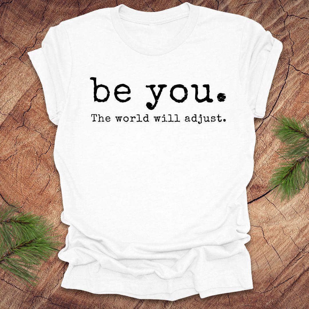 White t-shirt with 'be you. The world will adjust.' text on a wooden surface.