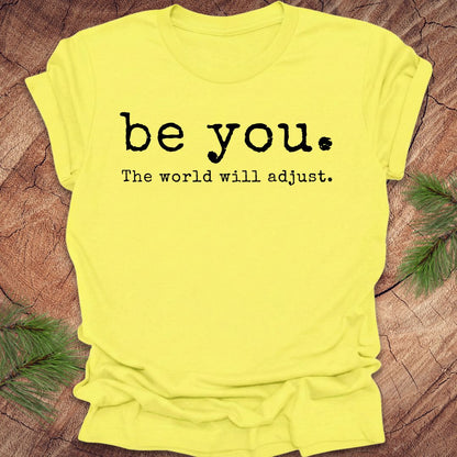 Yellow t-shirt with 'be you. The world will adjust.' text on a wooden surface.