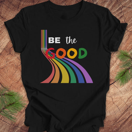 Black t-shirt with rainbow graphic and text, 'Be the Good' on a wood background.