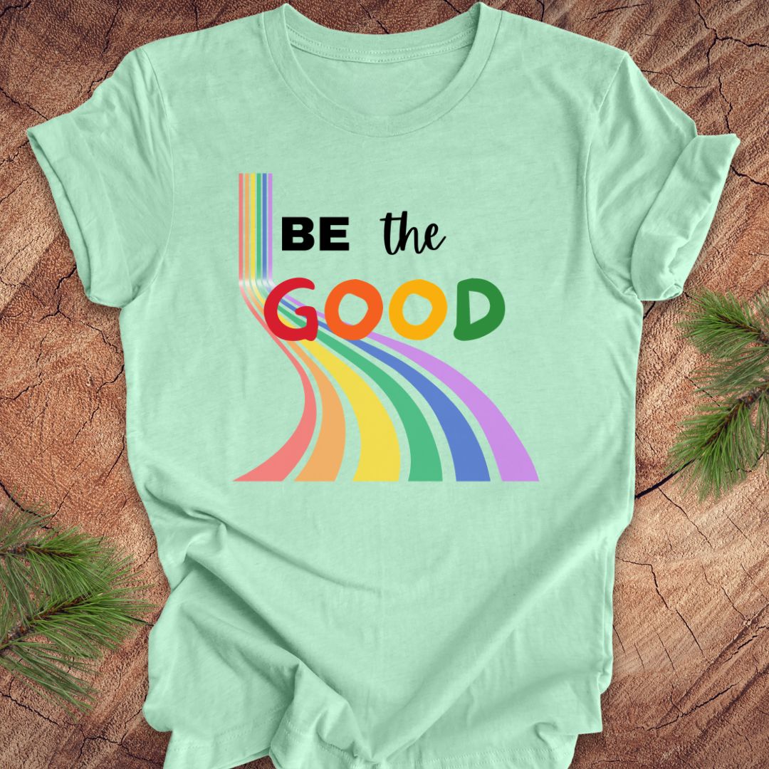 Mint green t-shirt with rainbow graphic and text, 'Be the Good' on a wood background.