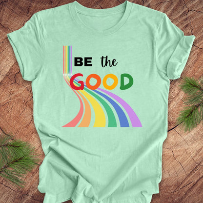 Mint green t-shirt with rainbow graphic and text, 'Be the Good' on a wood background.