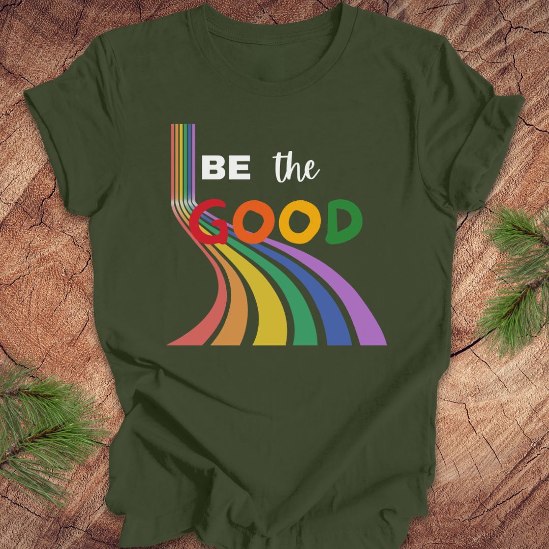 Green t-shirt with rainbow graphic and text, 'Be the Good' on a wood background.