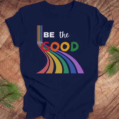 Navy blue t-shirt with rainbow graphic and text, 'Be the Good' on a wood background.
